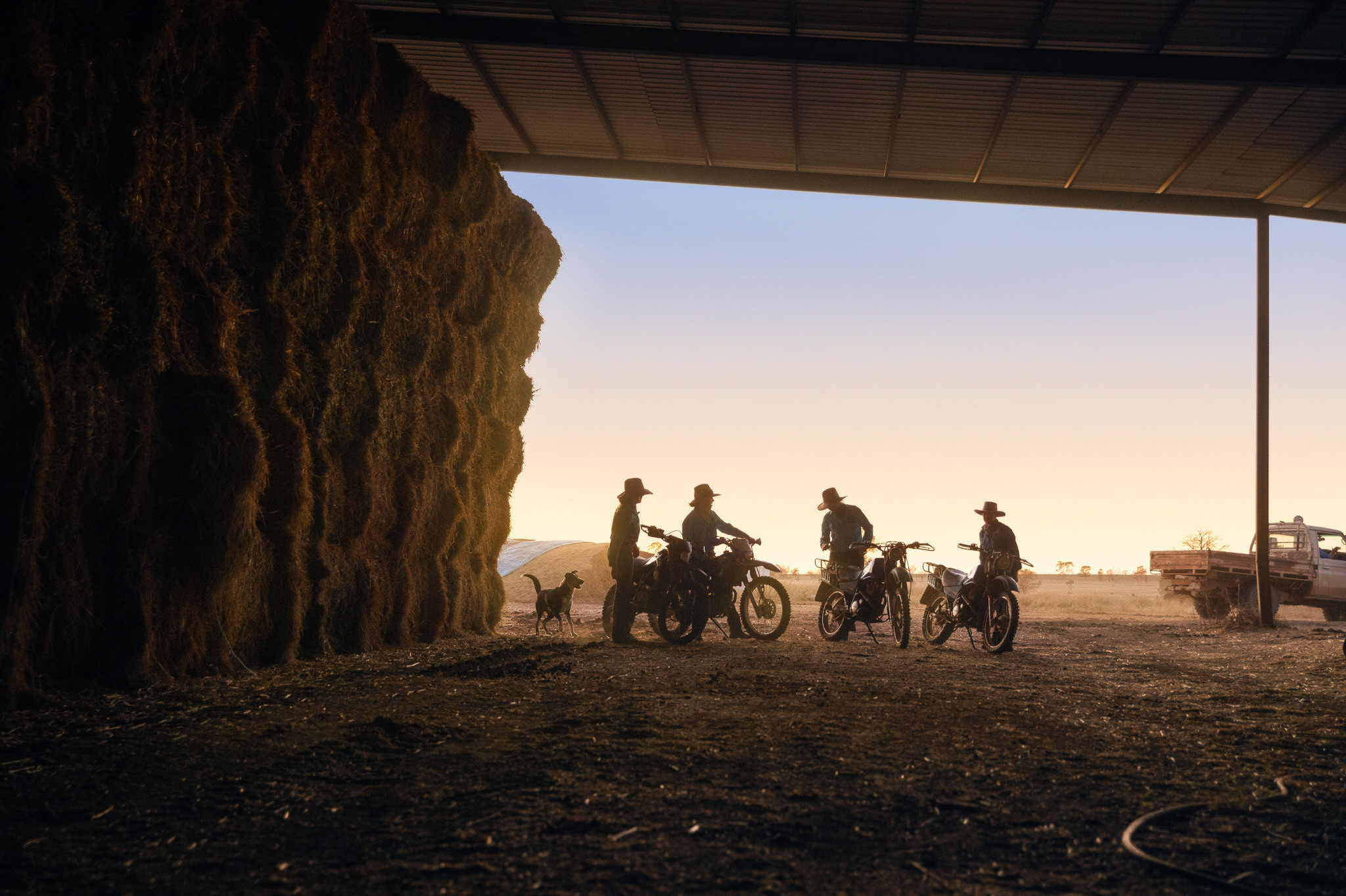 Five people on dirt bikes are silhouetted in a large open barn doorway with a large stack of hay bales during a vibrant sunset.