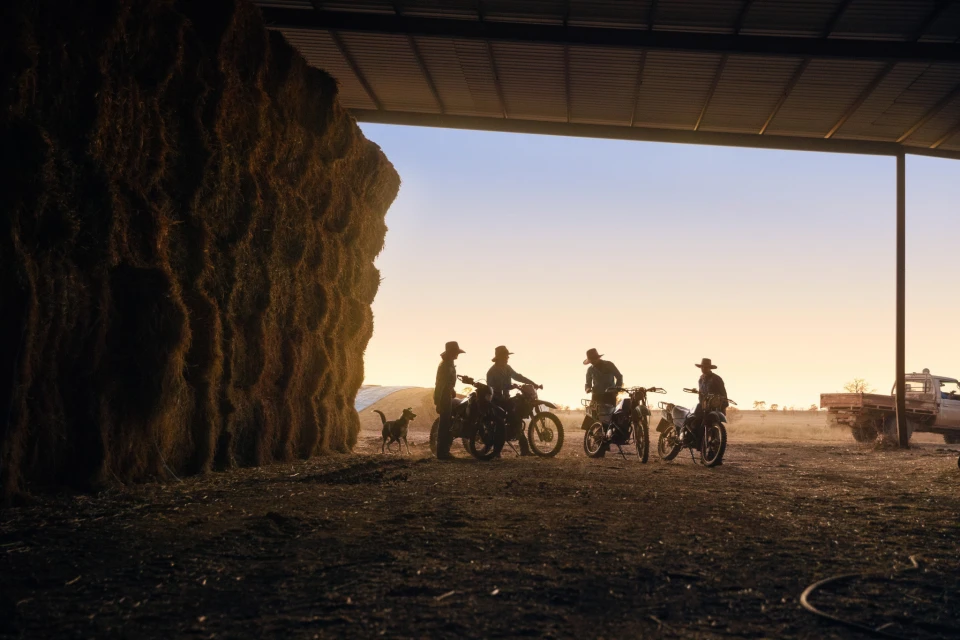 Five people on dirt bikes are silhouetted in a large open barn doorway with a large stack of hay bales during a vibrant sunset.