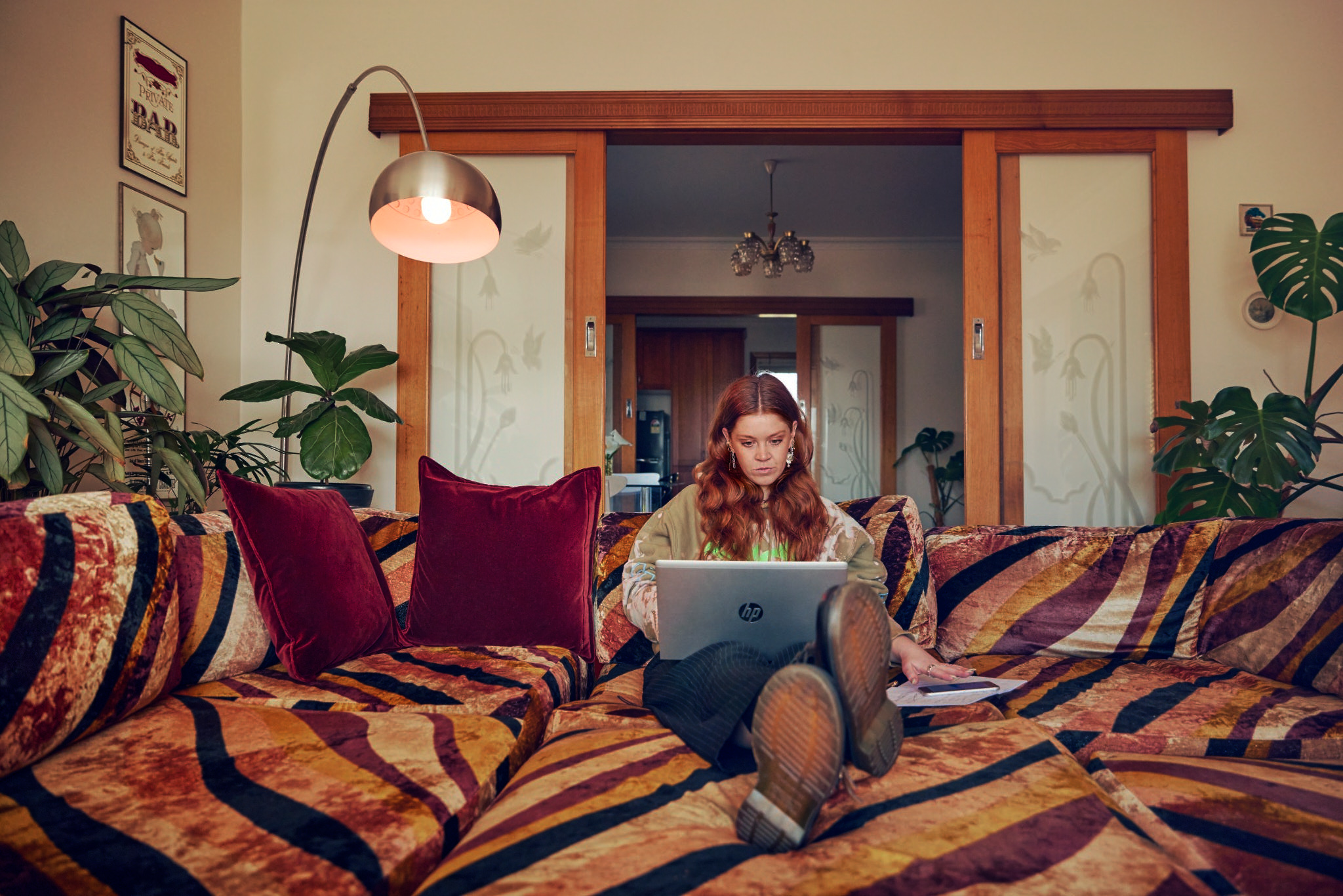 A woman with red hair sits on a tiger-striped couch and works on a laptop in a living room with indoor plants.