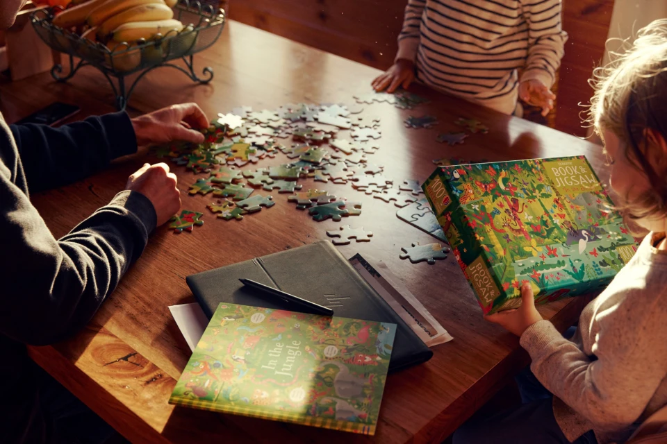 Two children and an adult completing a jigsaw puzzle on a wood table in a brightly lit interior with puzzle box and bananas.