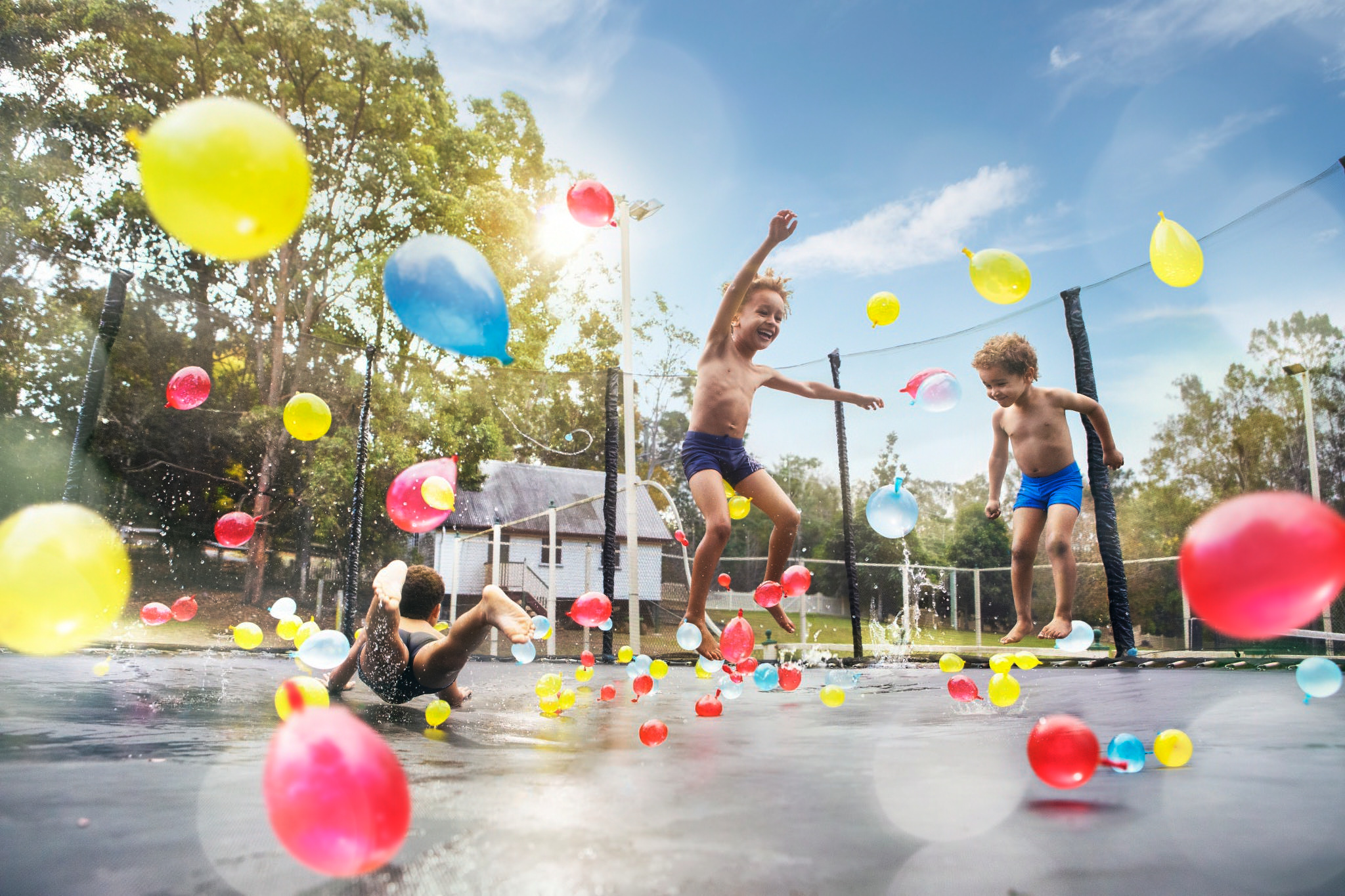 Three children play on a trampoline covered in water balloons under a blue sky with trees and a house in the background.