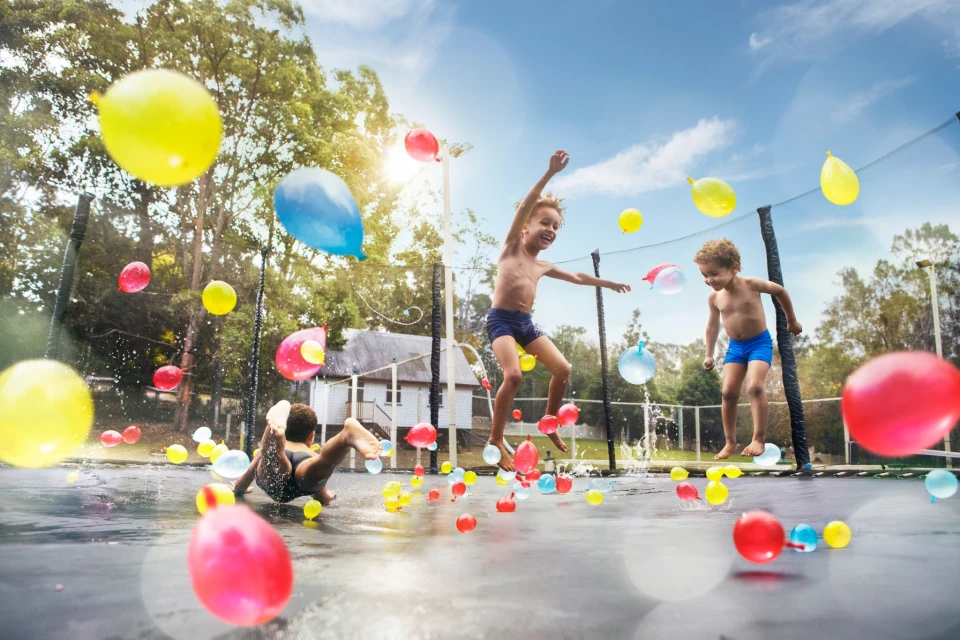 Three children play on a trampoline covered in water balloons under a blue sky with trees and a house in the background.