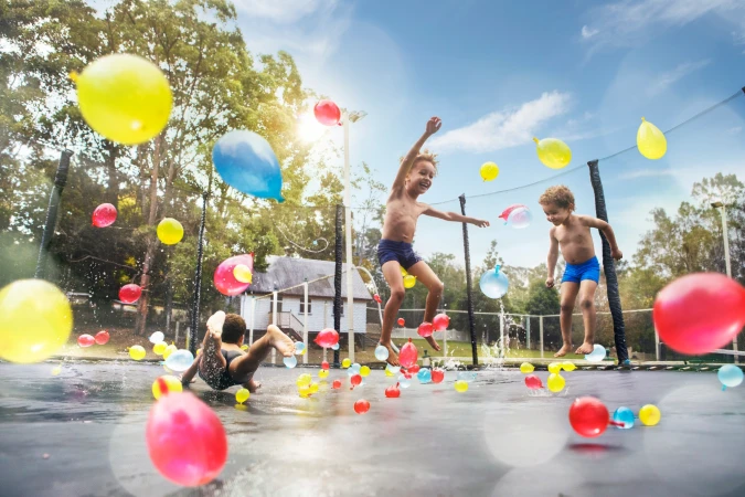 Three children play on a trampoline covered in water balloons under a blue sky with trees and a house in the background.