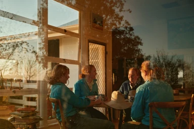 Four women sitting at a table together indoors, shot through a window reflection with warm golden light.