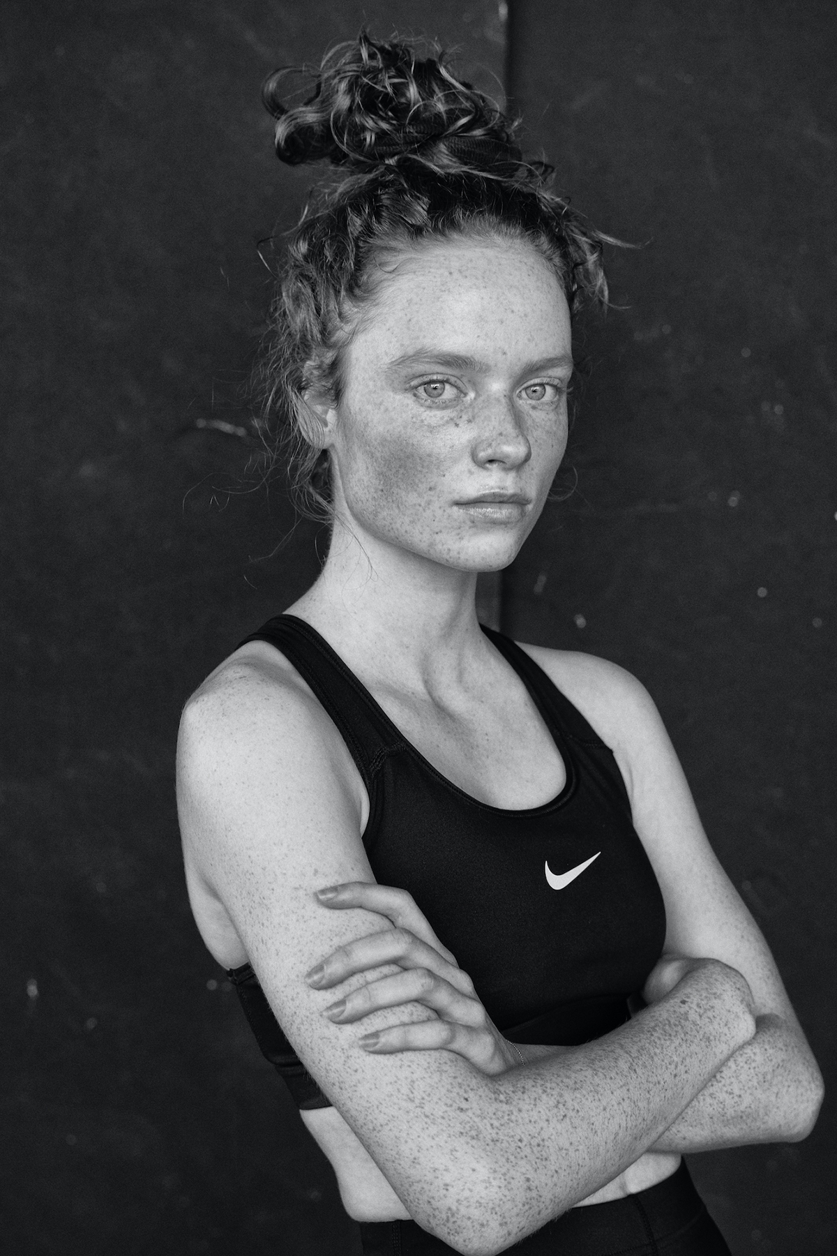 Monochrome portrait of a young adult woman with freckles and a messy bun, wearing a black sports bra and posing indoors.