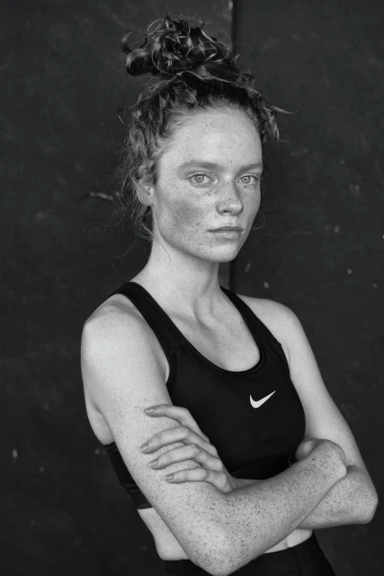 Monochrome portrait of a young adult woman with freckles and a messy bun, wearing a black sports bra and posing indoors.