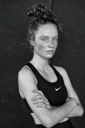 Monochrome portrait of a young adult woman with freckles and a messy bun, wearing a black sports bra and posing indoors.