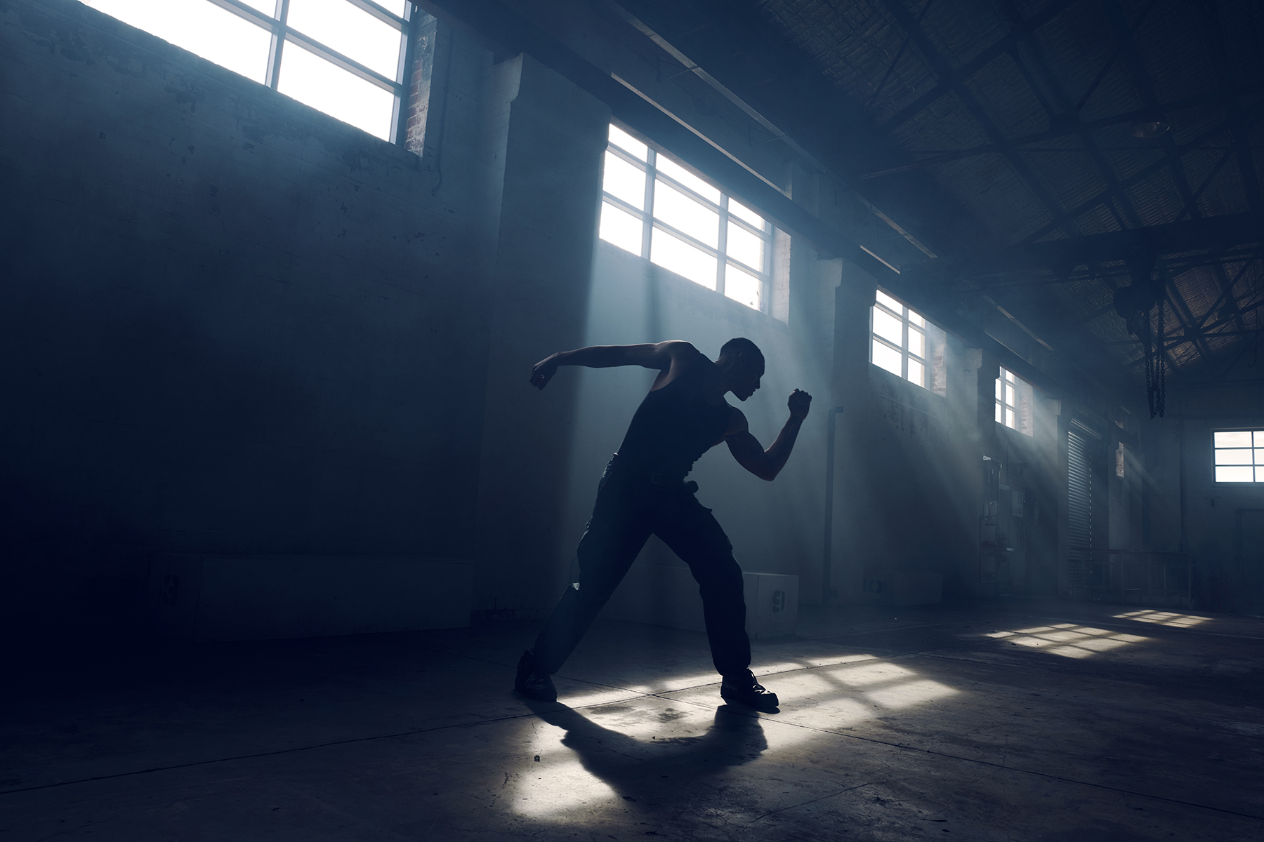 Silhouette of a muscular adult male in tank top and pants striking a dynamic pose in an empty warehouse lit by bright sunlight streaming through windows.