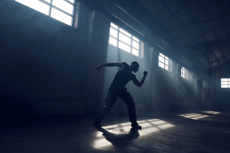 Silhouette of a muscular adult male in tank top and pants striking a dynamic pose in an empty warehouse lit by bright sunlight streaming through windows.
