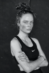 Black and white portrait of a freckled woman with hair in a high bun wearing a black sports bra with arms crossed
