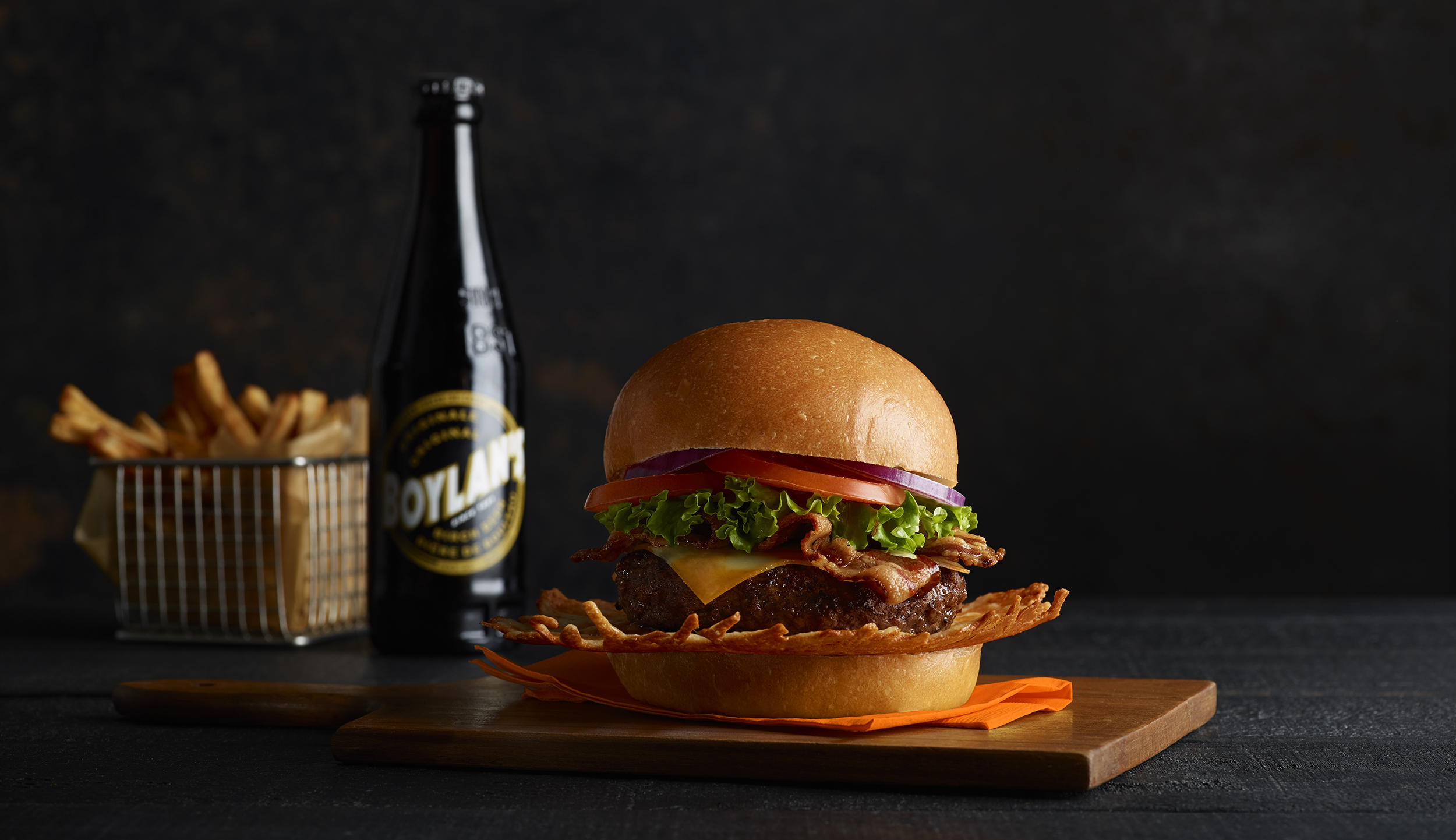 Studio shot of a gourmet burger with bacon, cheese, tomato, and lettuce on a wooden serving board next to a bottle of soda and fries.