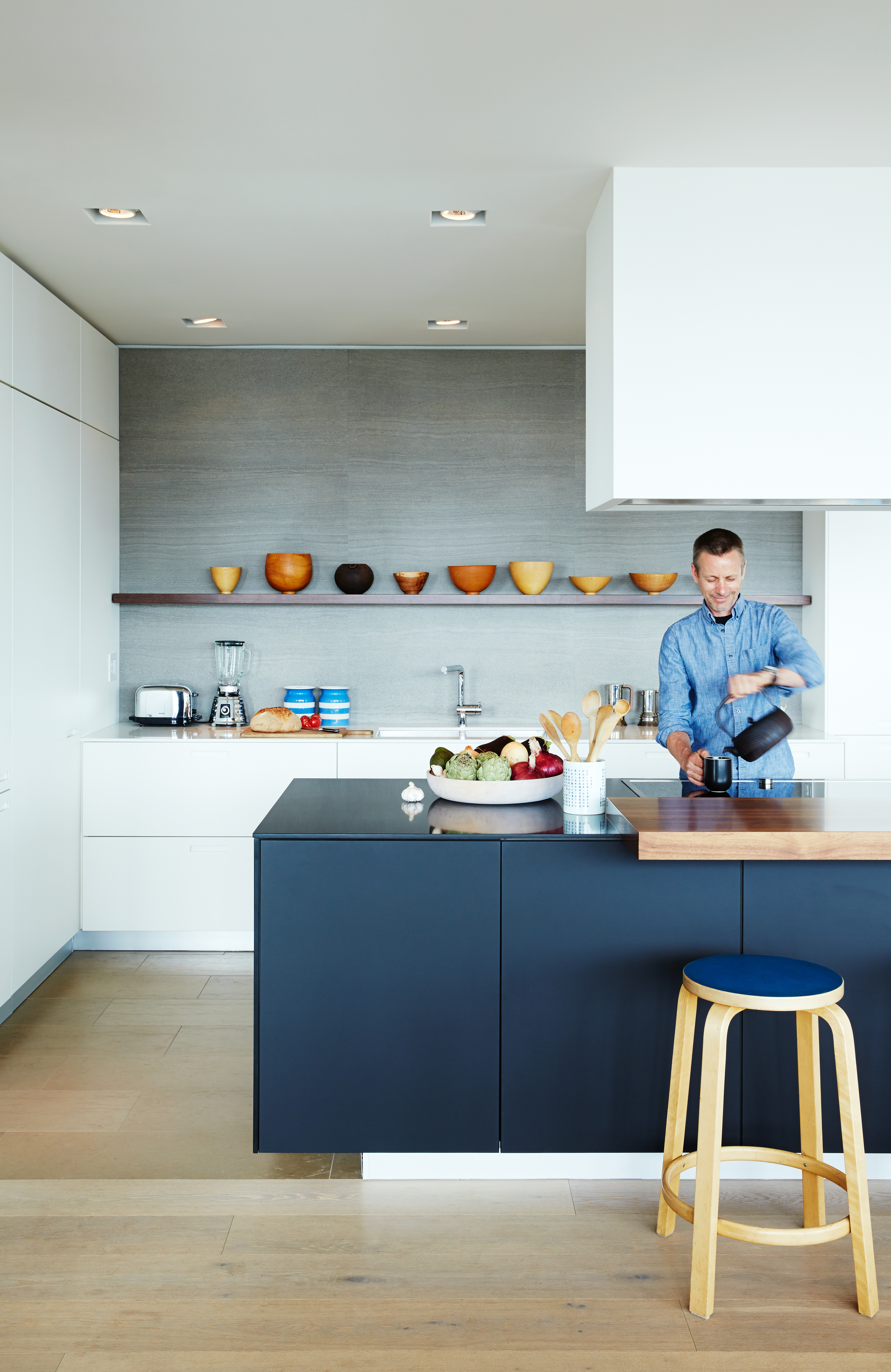 A man in a blue shirt pours liquid from a black kettle into a mug in a modern kitchen with white cabinets, dark island, and wooden stool.