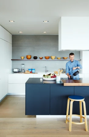 A man in a blue shirt pours liquid from a black kettle into a mug in a modern kitchen with white cabinets, dark island, and wooden stool.