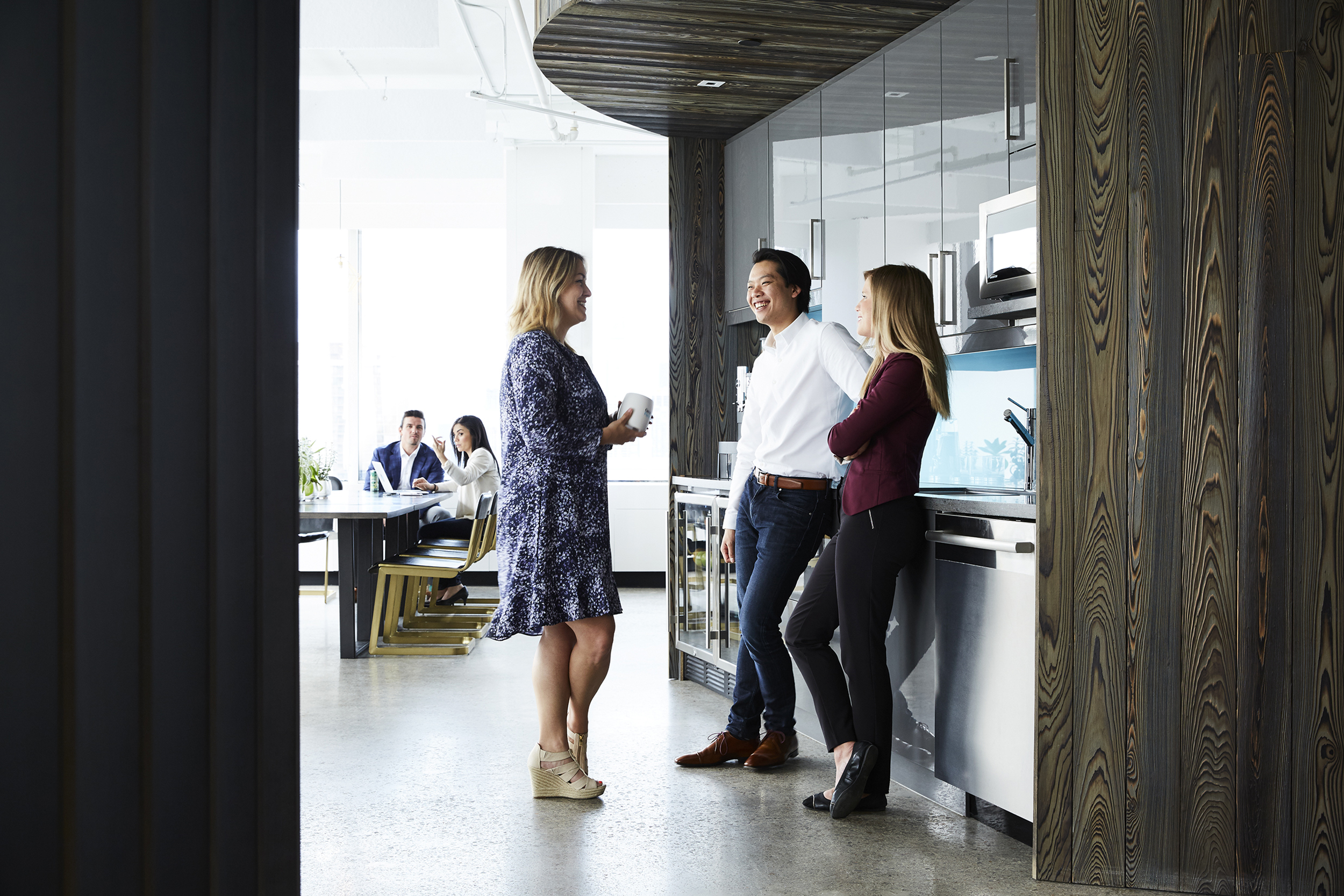 Three business people in casual office kitchen: woman in blue dress holds cup, man in white shirt smiles, woman in maroon leans on counter.