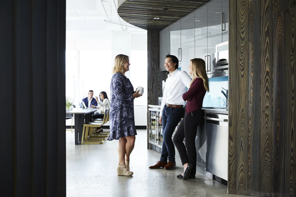Three business people in casual office kitchen: woman in blue dress holds cup, man in white shirt smiles, woman in maroon leans on counter.