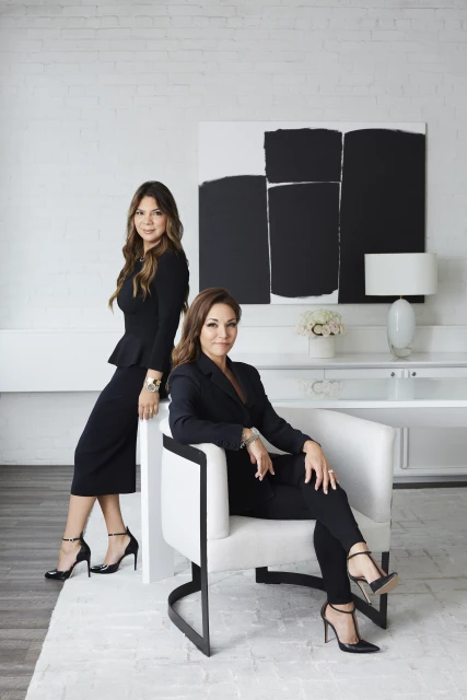 Two women in black business attire pose indoors; one sits in a white chair, the other stands behind her near an abstract painting.