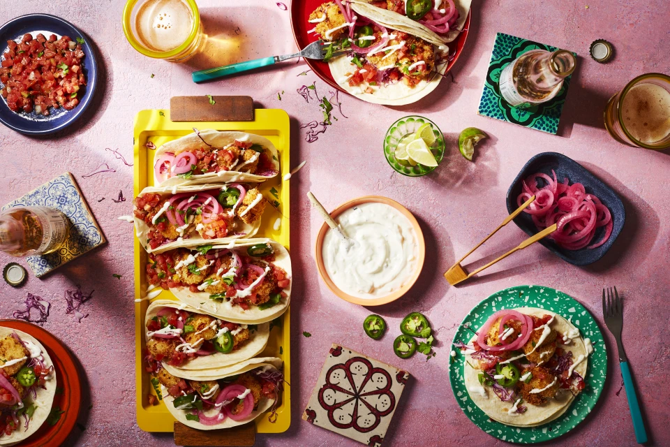 Overhead shot of fish tacos arranged on plates and a yellow tray with toppings and beer, set against a pink textured background.