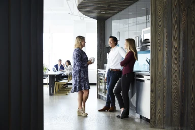 Three office workers talking in a modern kitchen space while two colleagues sit at a table in the background.