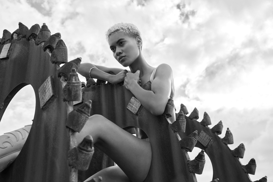 Black and white portrait of a young adult woman with short hair, posing on an abstract architectural metal structure against a cloudy sky.