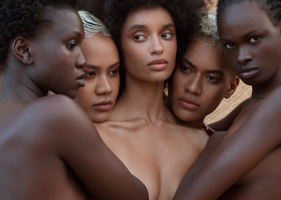 Artistic studio portrait of five adult women of diverse ethnicities closely posed together, softly lit against a textured background.