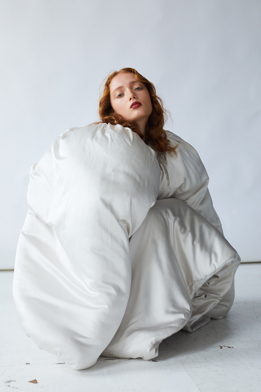 Portrait of a young woman with red hair crouching and wrapped in a puffy, off-white jacket against a light neutral background.