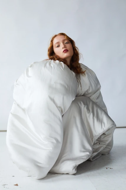 Portrait of a young woman with red hair crouching and wrapped in a puffy, off-white jacket against a light neutral background.