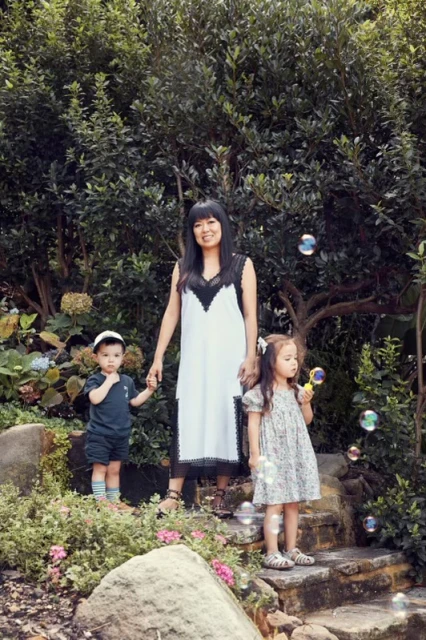 A mother stands in a garden with her two young children, who are playing with bubbles. Mature trees and flowering plants in the background.