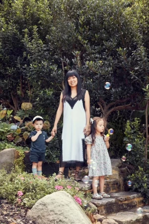 A mother stands in a garden with her two young children, who are playing with bubbles. Mature trees and flowering plants in the background.
