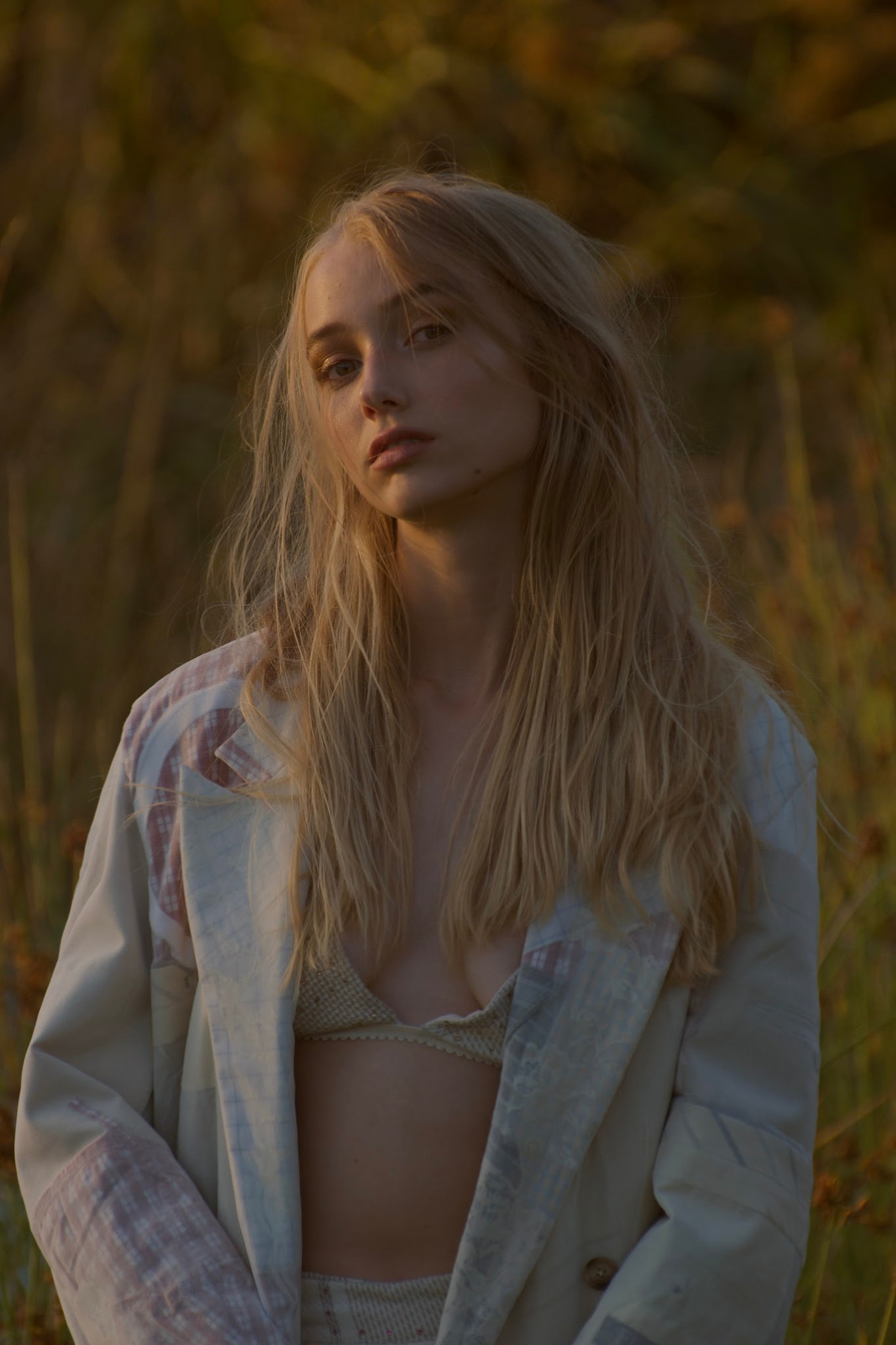 Portrait of a fair-skinned woman with long blonde hair wearing a patterned jacket, standing in a field with a natural, soft-focus background.