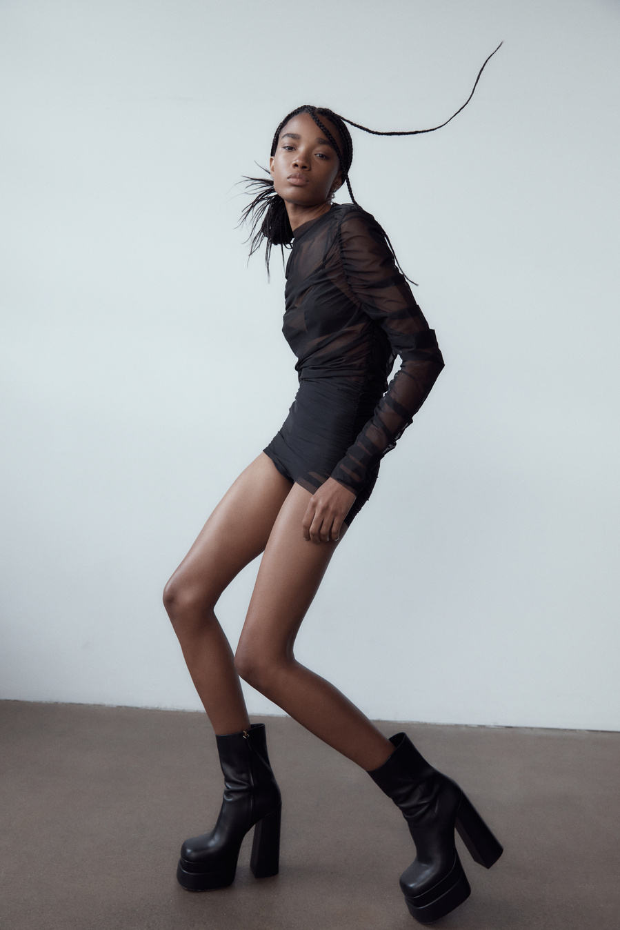 A slender, dark-skinned woman models a long-sleeved black dress with platform boots against a pale studio backdrop in a fashion shot.