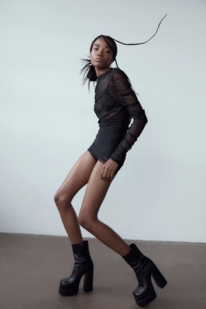 Black woman in a sheer mesh top and chunky platform boots posing against a neutral grey studio background