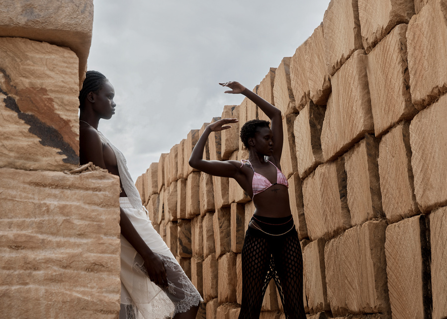Two dark-skinned women pose by stacked stones. One wears a white dress; the other wears a bikini top and pants against a neutral sky.