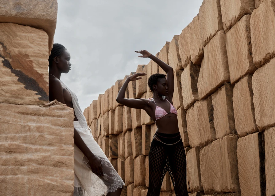 Two dark-skinned women pose by stacked stones. One wears a white dress; the other wears a bikini top and pants against a neutral sky.