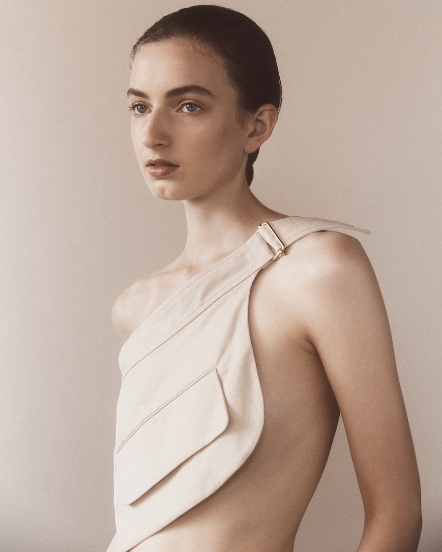 Close-up studio shot of a young woman wearing a beige asymmetrical top, gazing off-camera. The lighting is soft and diffused.