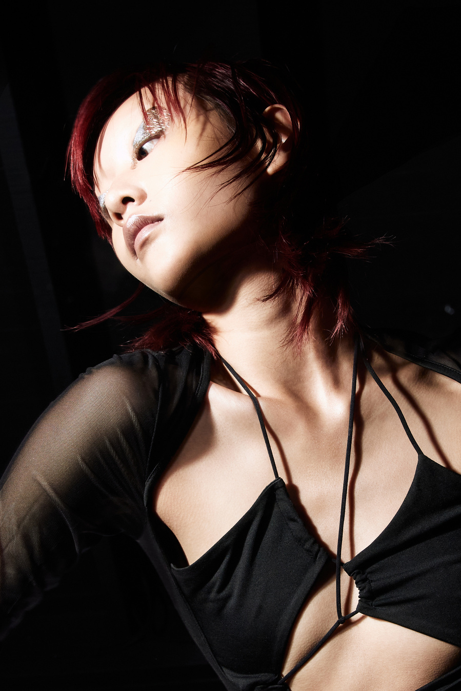 Close-up shot of a fashionable Asian adult female with red hair, metallic eye makeup, wearing a revealing black top in a dark studio.