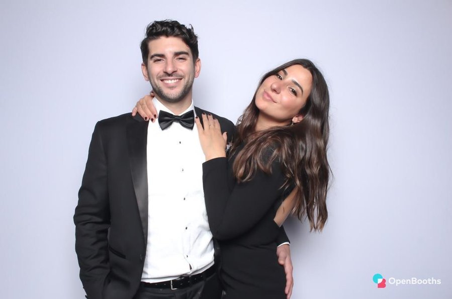Formal couple smiling in photo booth. Man in tuxedo with bow tie and woman in black dress with long sleeves and open back.