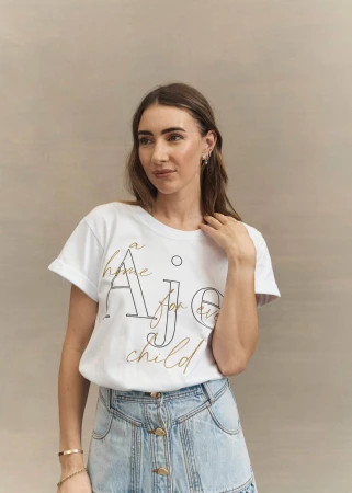 Woman wearing white graphic t-shirt and denim skirt with makeup by Lorella Giannini against a neutral textured wall.