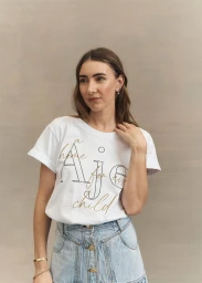Woman wearing white graphic t-shirt and denim skirt with makeup by Lorella Giannini against a neutral textured wall.