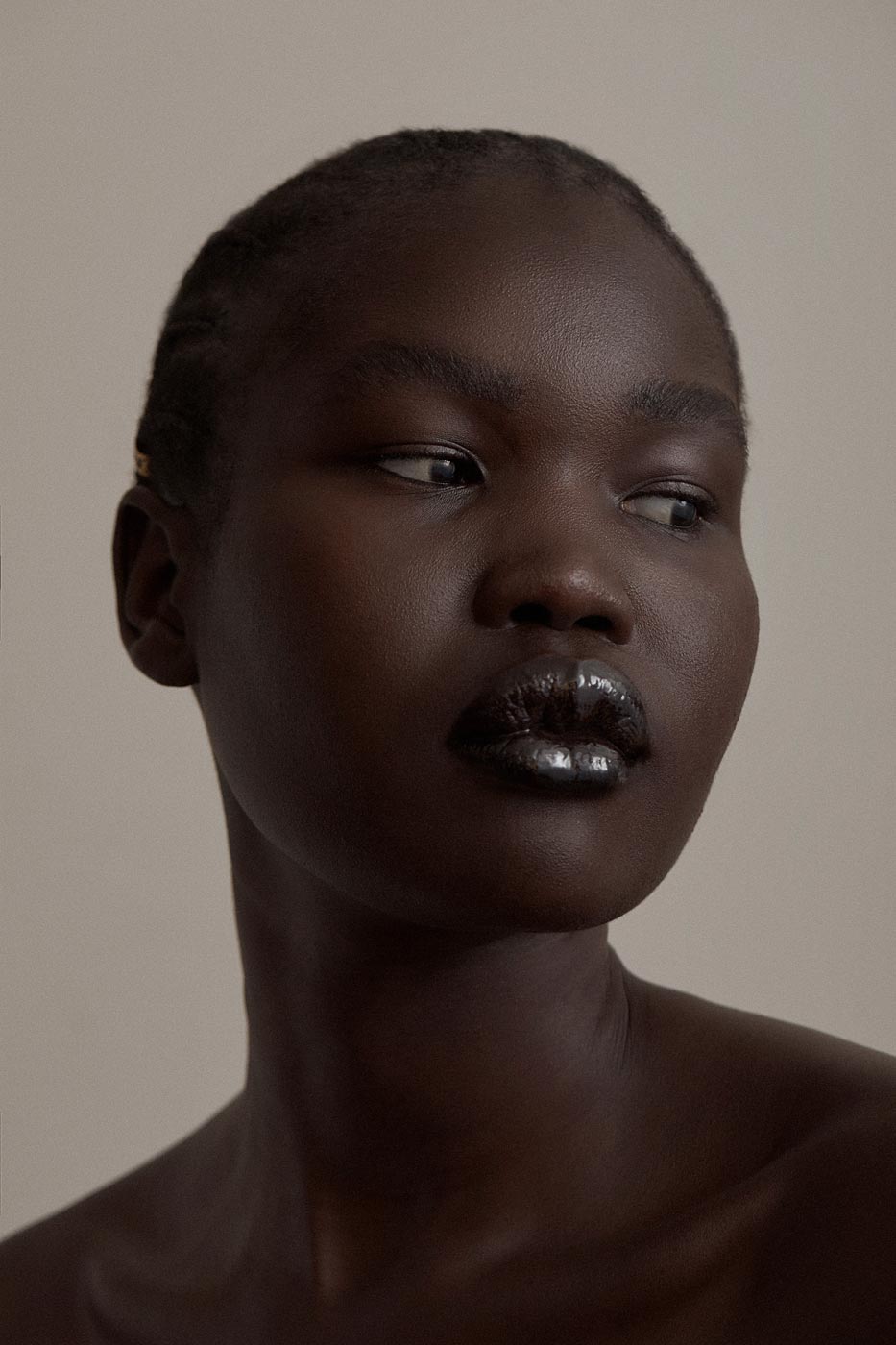 Close-up portrait of a woman with dark skin and glossy dark lips. She has a neutral expression and is set against a neutral background.