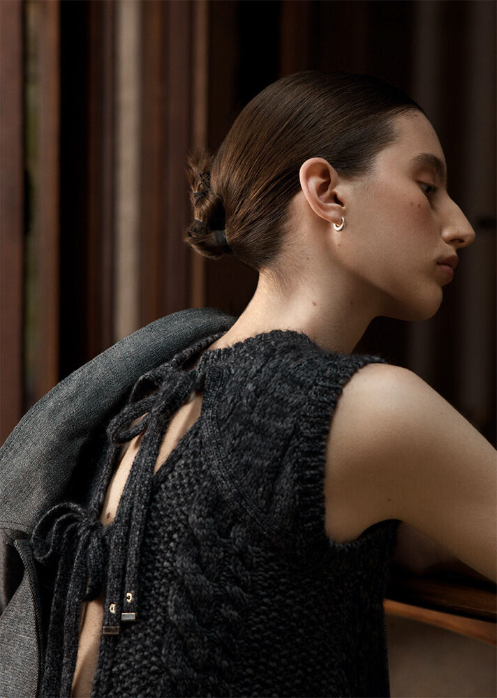 Portrait of a woman with an updo, wearing a dark knit top with an open back and ties, and a gray jacket over her shoulders.