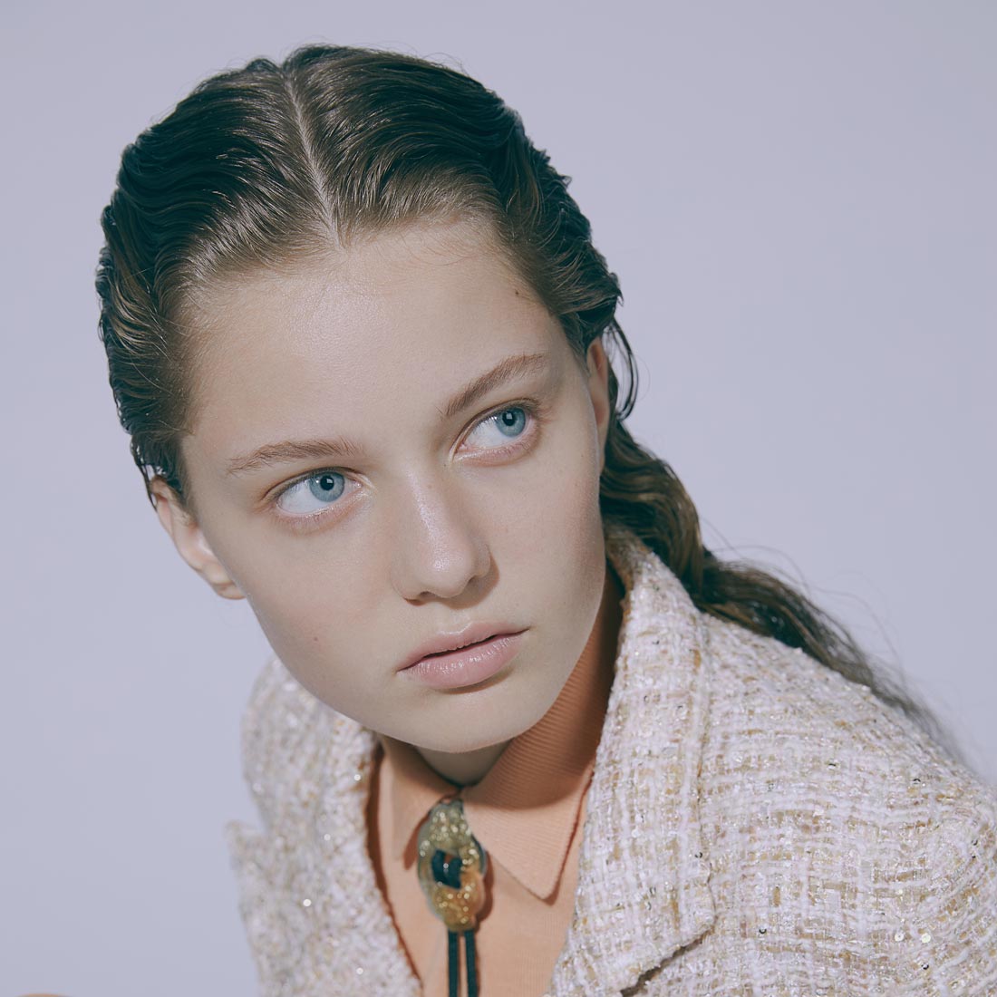 Portrait of a young woman with wavy brown hair and blue eyes wearing a cream tweed blazer and bolo tie, gazing to the side in a photo studio.