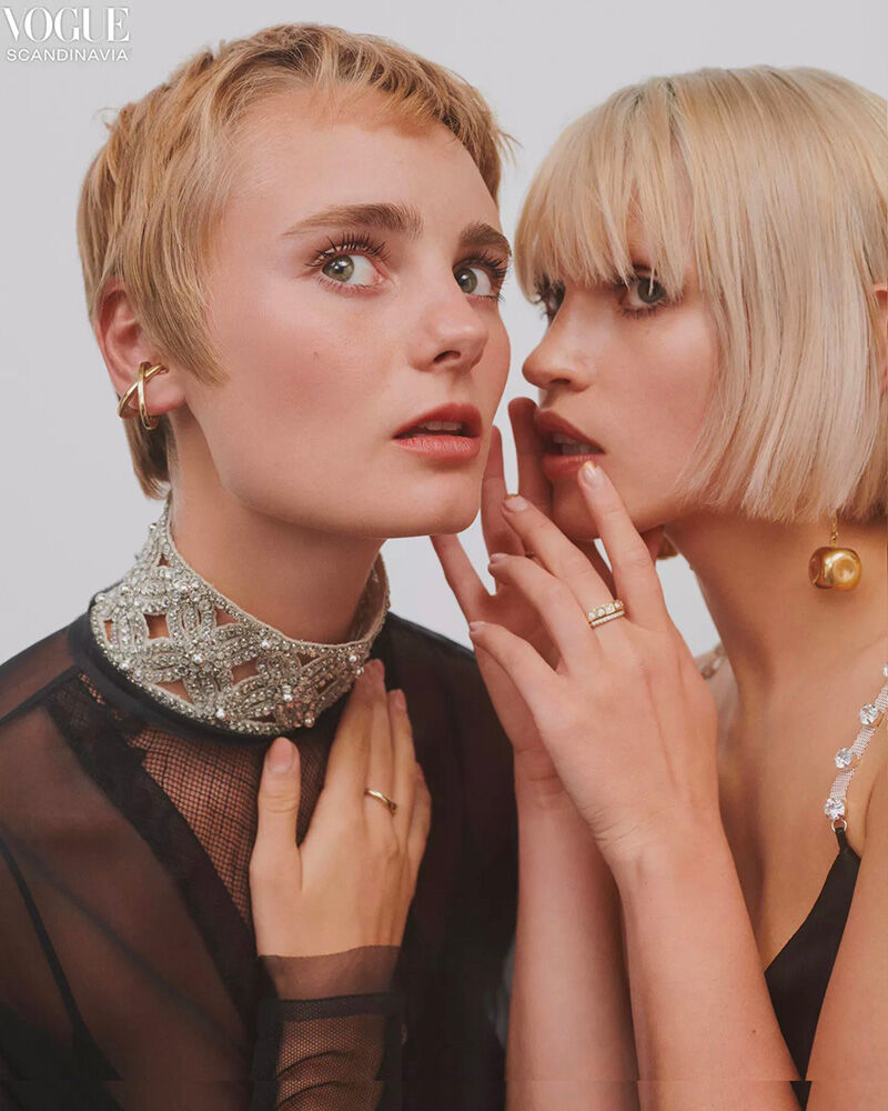 Two adult women pose indoors, one whispering with short blond hair, styled in a black dress with jeweled collar, in a fashion editorial.