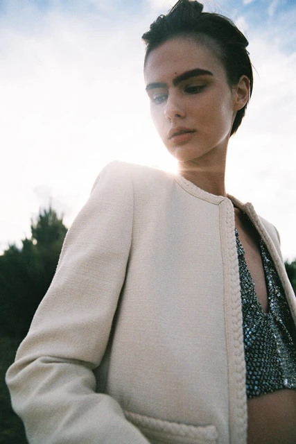 Outdoor fashion shot of a young adult woman in a cream blazer and sparkly top against a natural backdrop with a bright sky.