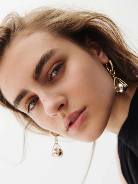 Close-up portrait of a young woman with fair skin and light brown hair, wearing gold earrings against a neutral backdrop.