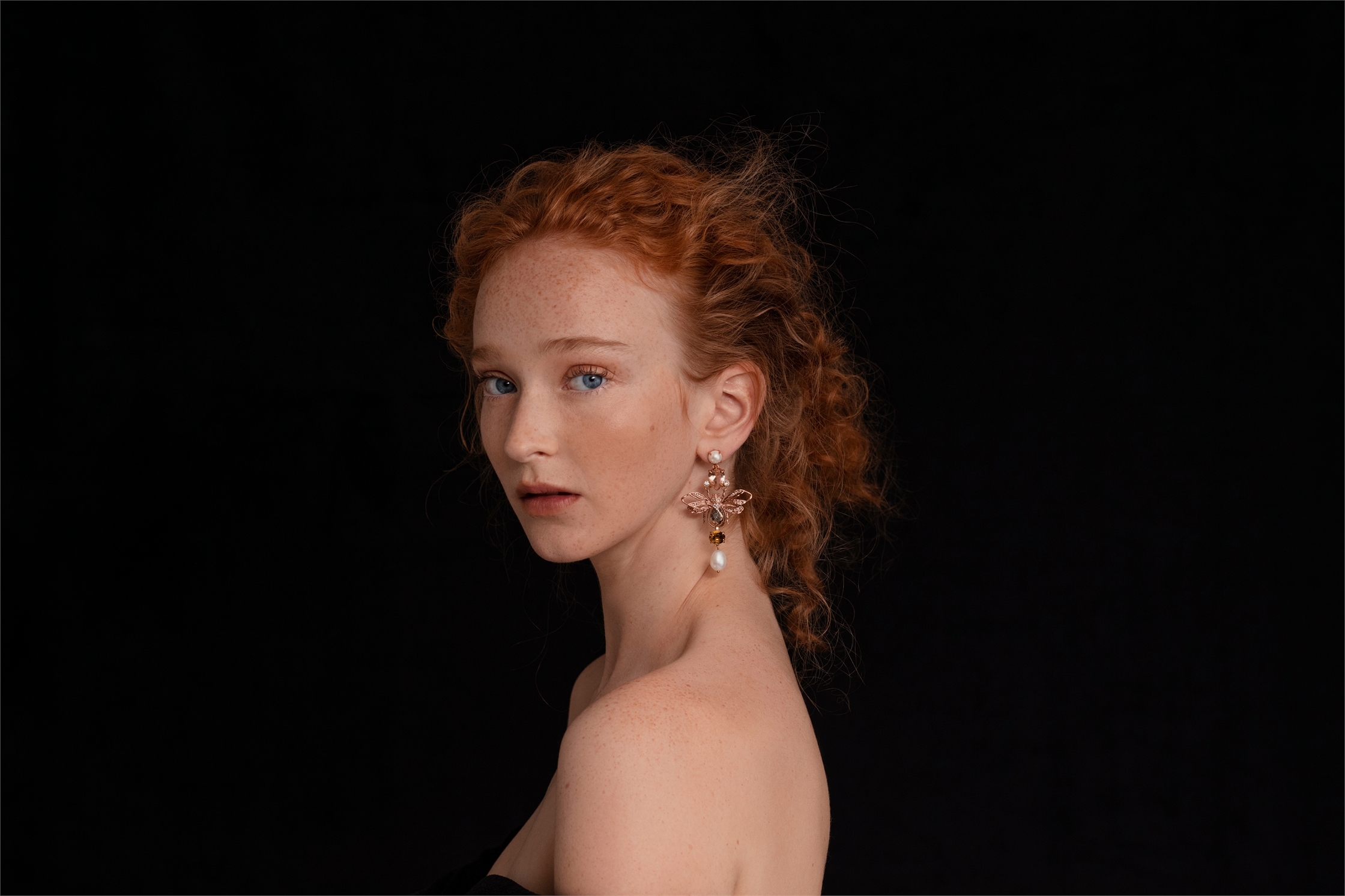 Close-up of a red-haired young woman with freckles wearing drop earrings against a dark background in a beauty fashion shot.