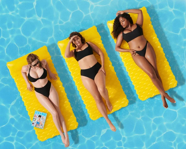 Three women in black swimsuits lounge on yellow inflatable rafts in a bright blue pool with rippling water on a sunny day.