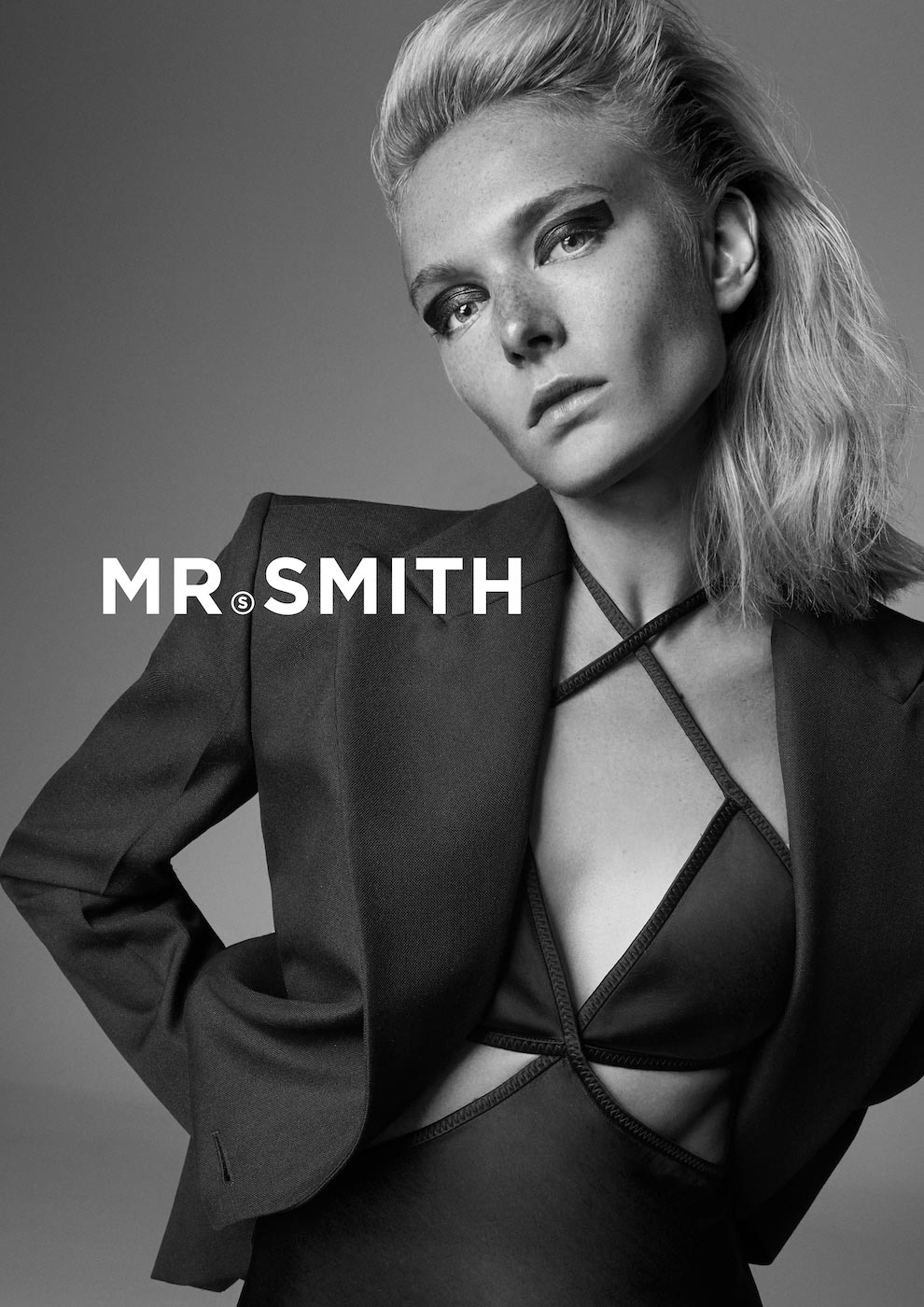 Black and white studio portrait of a blonde adult female model in a blazer and stylish eye makeup against a grey backdrop.