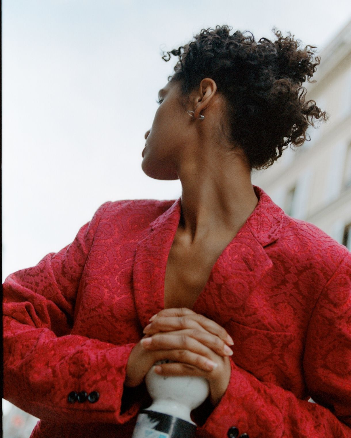 A woman with curly hair poses in a red patterned blazer outside against a light sky. The shot is a medium shot, taken from a low angle.