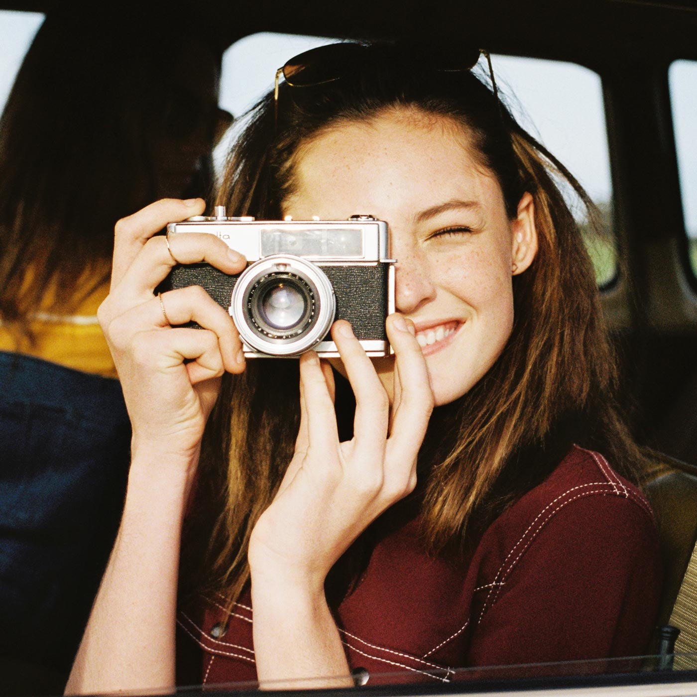 Young woman smiles as she looks through the viewfinder of a vintage camera, seated in a car with a passenger visible in the background.