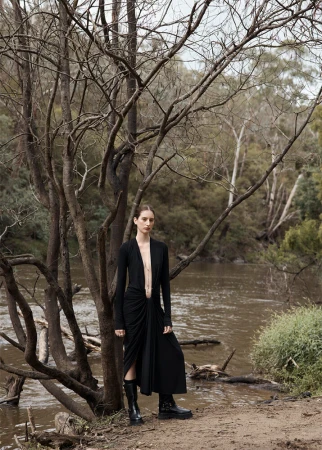 Model in a black dress and boots standing by a riverbank among bare trees, with make-up by Bradwyn Jones in Sydney.